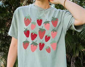 Puede incluir: Camiseta verde salvia con un estampado de fresas rojas y rosas con tallos verdes. Las fresas están dispuestas en un diseño de cuadrícula. La camiseta es de estilo casual y cómodo.