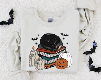 Puede incluir: Una sudadera de color crema con un gato negro durmiendo sobre una pila de libros. El gato está rodeado de velas y una calabaza tallada. Hay murciélagos volando alrededor del gato.