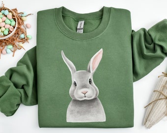Può includere: Una felpa verde con un'illustrazione di un coniglio grigio sul davanti. Il coniglio guarda direttamente lo spettatore. La felpa è a collo rotondo.