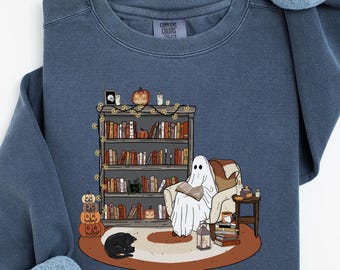 May include: A blue sweatshirt featuring a cozy Halloween scene. A ghost reads in a chair near a bookshelf, with a black cat, pumpkins, and a lantern. The sweatshirt has long sleeves and a crew neck.