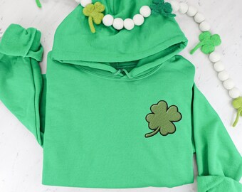 May include: A green hooded sweatshirt with a four-leaf clover embroidered on the front. The sweatshirt is folded and laid flat on a white surface.