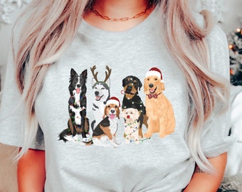 Puede incluir: Una camiseta gris con un gráfico de cinco perros con gorros de Navidad y luces. Los perros son un border collie blanco y negro, un husky con cuernos de reno, un beagle, un rottweiler y un golden retriever. El texto "EMMA & ANNA" está impreso en la parte inferior de la camiseta.