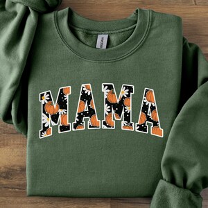 Op de afbeelding: Een olijfgroene sweatshirt met het woord "MAMA" in een witte, gebogen letter. De letters zijn gevuld met een patroon van oranje pompoenen en witte madeliefjes op een zwarte achtergrond. De sweatshirt heeft lange mouwen.