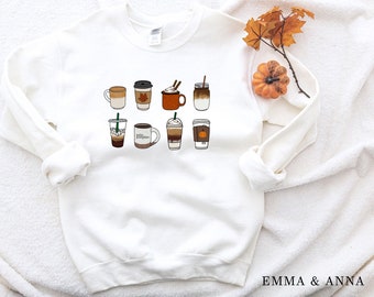May include: White sweatshirt with a graphic of various coffee drinks, including lattes, iced coffees, and pumpkin spice lattes. The text "Emma & Anna" is printed on the bottom right corner of the sweatshirt.