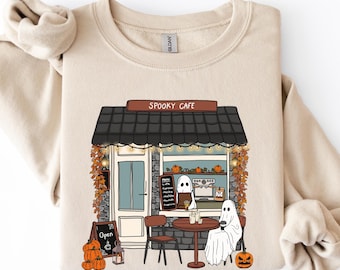 May include: A beige crewneck sweatshirt featuring a graphic illustration of a storefront labeled SPOOKY CAFE. The design depicts two white ghosts sitting at a small round table outside, surrounded by orange pumpkins and autumn foliage.