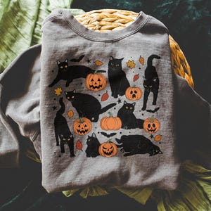 May include: A gray sweatshirt featuring a Halloween-themed design. The graphic includes black cats, orange pumpkins with carved faces, and autumn leaves. The cats are in various poses, creating a playful and festive look.