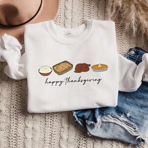 K&ouml;nnte beinhalten: Wei&szlig;es Sweatshirt mit einem Thanksgiving-Motiv mit einem Truthahn, Kartoffelp&uuml;ree, F&uuml;llung und K&uuml;rbiskuchen. Der Text "happy thanksgiving" ist unter den Speisen gedruckt.