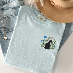 May include: A light blue t-shirt with a black cat embroidered on the front, surrounded by flowers. The shirt is folded next to a pair of light blue jeans, a tan hat, and a gold necklace.