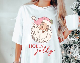 May include: White oversized t-shirt featuring a pink and white cartoon Santa Claus graphic and the text "HOLLY jolly" in red.