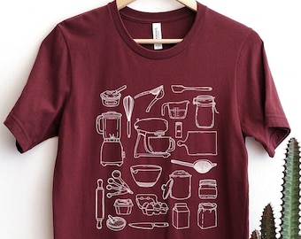 May include: A maroon t-shirt with a white outline of various kitchen tools and appliances. The design includes a blender, a mixer, a whisk, a rolling pin, a measuring cup, a measuring spoon, a spatula, a knife, a jar, a bowl, and a box of eggs.