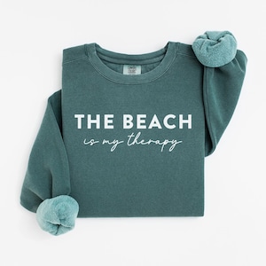 Pu&ograve; includere: Una felpa color teal con la scritta bianca "THE BEACH is my therapy". Le maniche sono arrotolate. Il tessuto sembra morbido. &Egrave; un capo d'abbigliamento casual.
