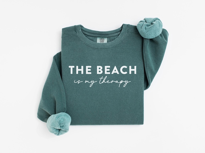 Pu&ograve; includere: Una felpa color teal con la scritta bianca "THE BEACH is my therapy". Le maniche sono arrotolate. Il tessuto sembra morbido. &Egrave; un capo d'abbigliamento casual.