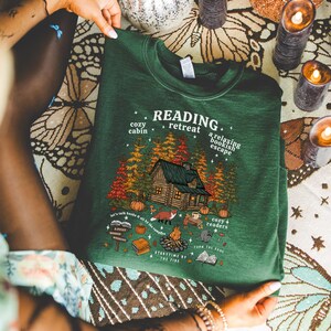 Op de afbeelding: Een donkergroene sweatshirt met de tekst "READING retreat" en een huttenafbeelding. De afbeelding bevat bomen, een vos, een kampvuur en de tekst "cozy cabin" en "a relaxing bookish escape".