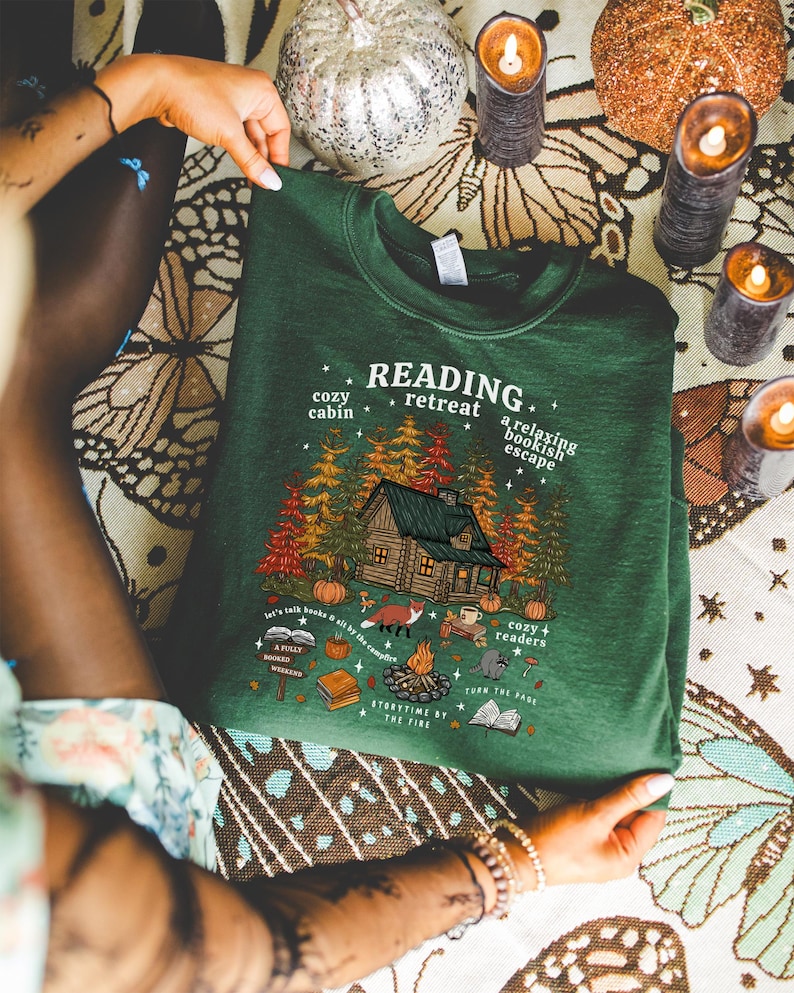 Op de afbeelding: Een donkergroene sweatshirt met de tekst "READING retreat" en een huttenafbeelding. De afbeelding bevat bomen, een vos, een kampvuur en de tekst "cozy cabin" en "a relaxing bookish escape".