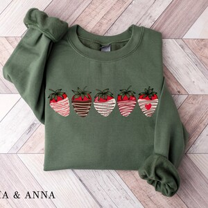 Puede incluir: Una sudadera verde con un gr&aacute;fico de cinco fresas cubiertas de chocolate. Las fresas son rojas con rayas blancas y marrones, y una tiene un peque&ntilde;o coraz&oacute;n rojo.