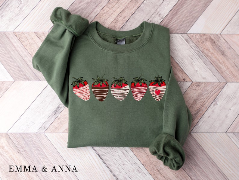 Puede incluir: Una sudadera verde con un gr&aacute;fico de cinco fresas cubiertas de chocolate. Las fresas son rojas con rayas blancas y marrones, y una tiene un peque&ntilde;o coraz&oacute;n rojo.