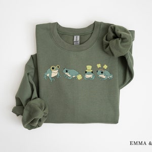 Puede incluir: Una sudadera verde con un gr&aacute;fico de cuatro ranas que llevan sombreros y gafas. Las ranas est&aacute;n rodeadas de tr&eacute;boles. La sudadera tiene cuello redondo y mangas largas.