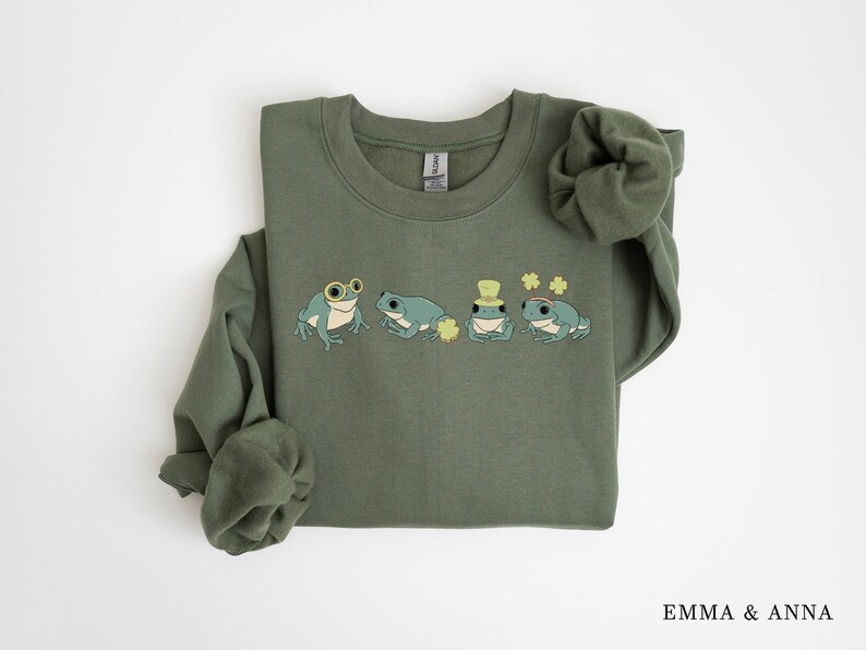 Puede incluir: Una sudadera verde con un gr&aacute;fico de cuatro ranas que llevan sombreros y gafas. Las ranas est&aacute;n rodeadas de tr&eacute;boles. La sudadera tiene cuello redondo y mangas largas.