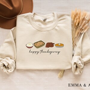 May include: Cream-colored sweatshirt with a Thanksgiving-themed graphic. The design includes illustrations of mashed potatoes, stuffing, a roasted turkey, and pumpkin pie, with the words "happy thanksgiving" in black script. A brown hat and dried wheat are in the background.