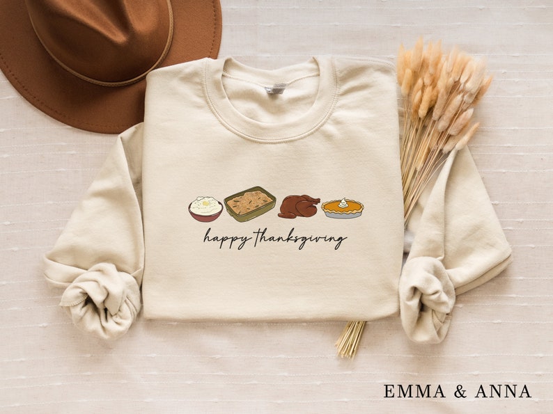 May include: Cream-colored sweatshirt with a Thanksgiving-themed graphic. The design includes illustrations of mashed potatoes, stuffing, a roasted turkey, and pumpkin pie, with the words "happy thanksgiving" in black script. A brown hat and dried wheat are in the background.