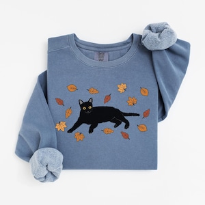 May include: A blue-grey sweatshirt featuring a black cat illustration with yellow eyes, surrounded by falling autumn leaves in shades of orange and brown. The sweatshirt has a crew neck and long sleeves with a scrunchie on the sleeve.