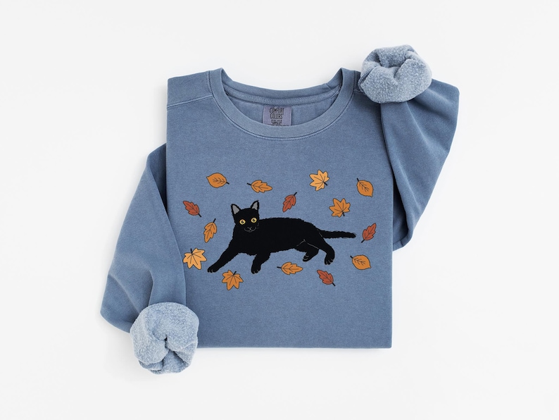 May include: A blue-grey sweatshirt featuring a black cat illustration with yellow eyes, surrounded by falling autumn leaves in shades of orange and brown. The sweatshirt has a crew neck and long sleeves with a scrunchie on the sleeve.