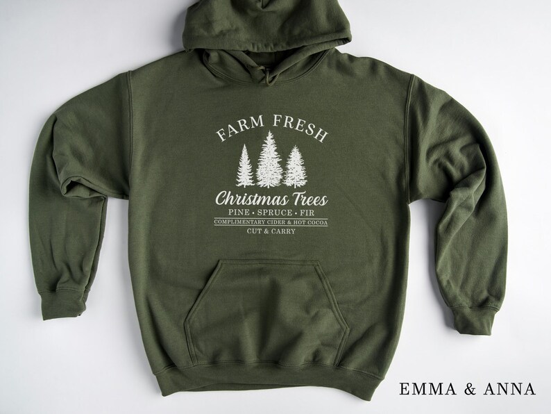 Op de afbeelding: Een groene hoodie met de tekst "FARM FRESH Christmas Trees PINE SPRUCE FIR COMPLIMENTARY CIDER & HOT COCOA CUT & CARRY" en een afbeelding van drie gestileerde dennenbomen.