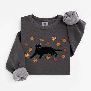 May include: Dark grey sweatshirt featuring a black cat illustration with yellow eyes, surrounded by autumn leaves in shades of orange and brown. The long-sleeved shirt has a crew neck and ribbed cuffs.