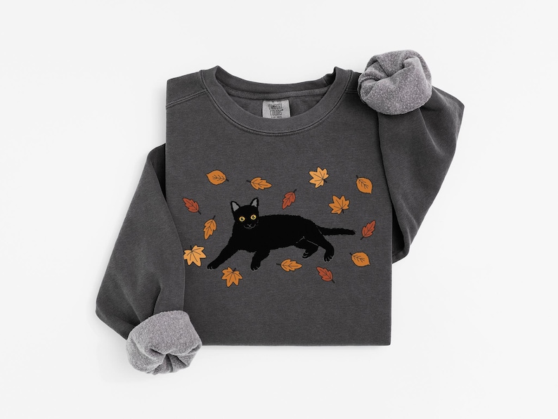 May include: Dark grey sweatshirt featuring a black cat illustration with yellow eyes, surrounded by autumn leaves in shades of orange and brown. The long-sleeved shirt has a crew neck and ribbed cuffs.