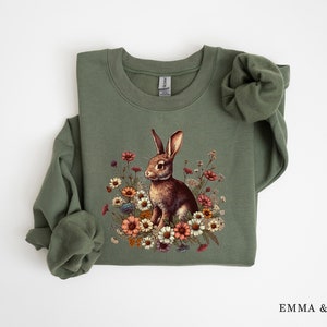 Pu&ograve; includere: Una felpa verde oliva con una dettagliata illustrazione di un coniglio marrone circondato da fiori colorati. Il design include fiori nei toni del rosa, arancione e bianco. La felpa ha maniche lunghe e girocollo.