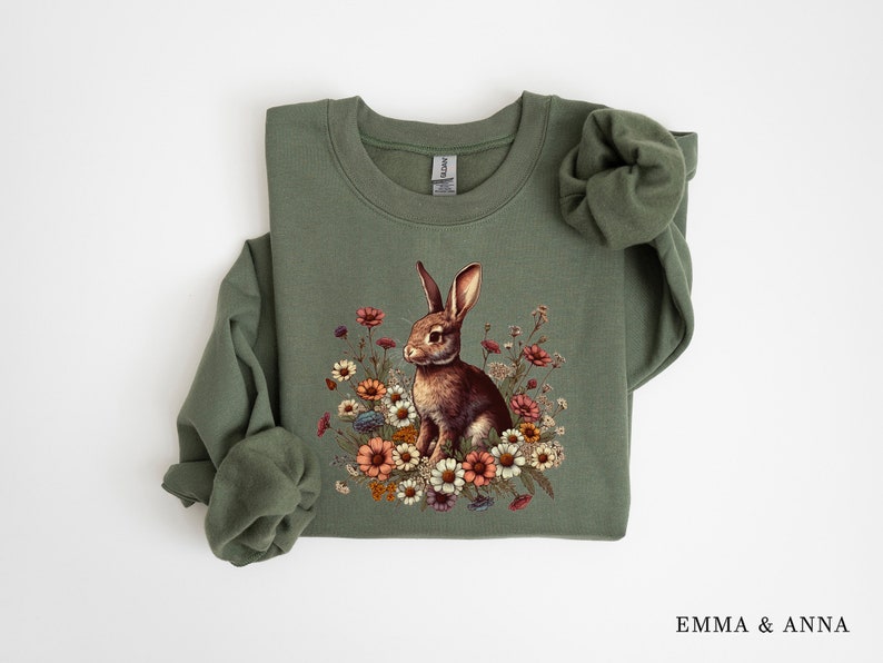 Pu&ograve; includere: Una felpa verde oliva con una dettagliata illustrazione di un coniglio marrone circondato da fiori colorati. Il design include fiori nei toni del rosa, arancione e bianco. La felpa ha maniche lunghe e girocollo.