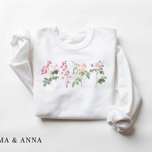 May include: White sweatshirt with a floral print of four different flowers. The flowers are pink, white, and peach. The text "EMMA & ANNA" is printed below the flowers.