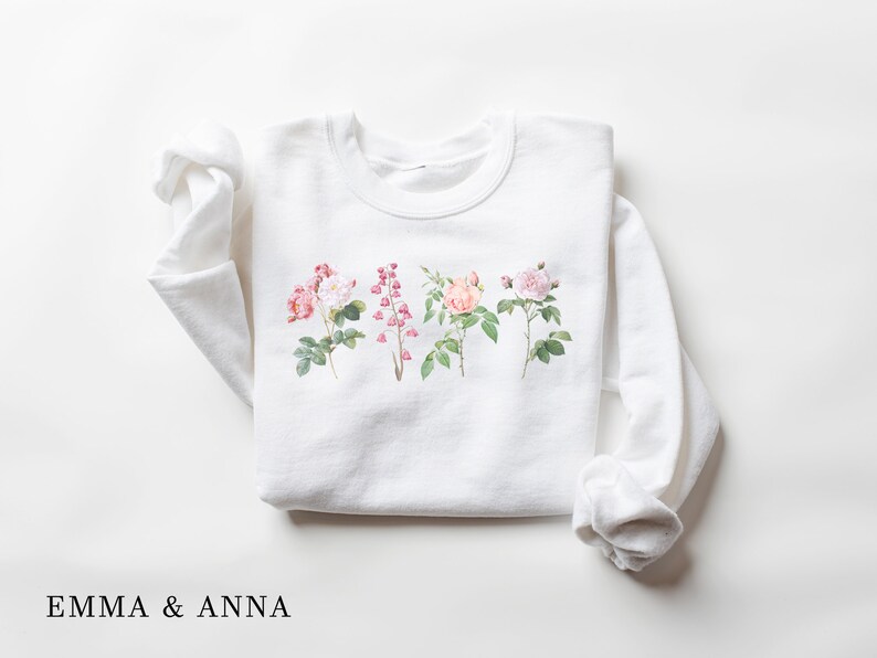 May include: White sweatshirt with a floral print of four different flowers. The flowers are pink, white, and peach. The text "EMMA & ANNA" is printed below the flowers.