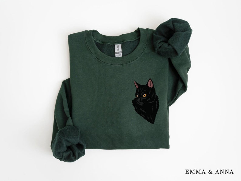 May include: A dark green sweatshirt with a black cat graphic on the front. The cat has yellow eyes and a pink nose.