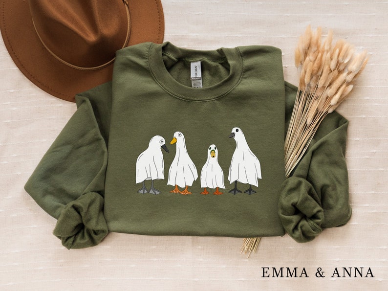 May include: A green sweatshirt with a graphic of four ghost ducks. The ducks are white with black eyes and orange beaks. The text "EMMA & ANNA" is printed on the bottom of the sweatshirt.