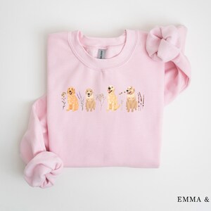 May include: Pink sweatshirt with a graphic of four golden retriever dogs sitting in a row with flowers and greenery behind them.