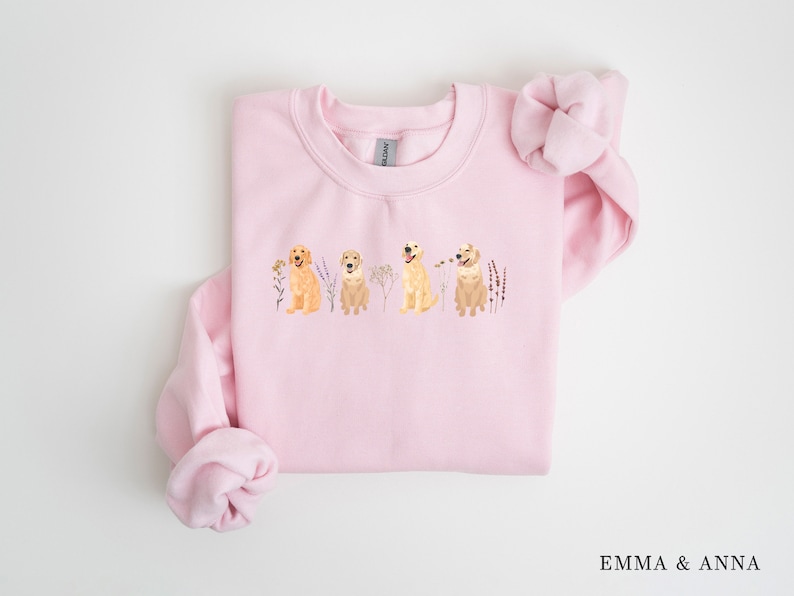 May include: Pink sweatshirt with a graphic of four golden retriever dogs sitting in a row with flowers and greenery behind them.