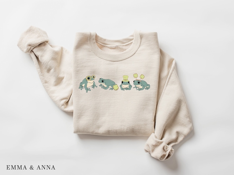 Puede incluir: Una sudadera beige con un gr&aacute;fico de cuatro ranas con sombreros y sosteniendo tr&eacute;boles. Las ranas son verdes con ojos negros y vientres blancos.