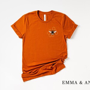 May include: A rust-colored t-shirt with a bee and honeycomb design on the front. The text "EMMA & ANNA" is printed on the bottom of the image.