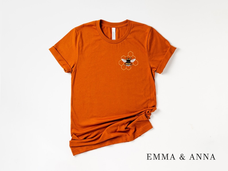 May include: A rust-colored t-shirt with a bee and honeycomb design on the front. The text "EMMA & ANNA" is printed on the bottom of the image.