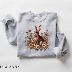 Pu&ograve; includere: Felpa grigio chiaro con un disegno di un coniglio marrone circondato da fiori colorati. Il coniglio ha lunghe orecchie ed &egrave; seduto. Il testo "EMMA & ANNA" &egrave; stampato in basso.