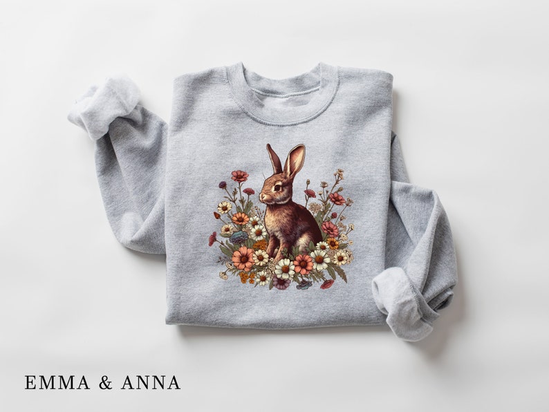 Pu&ograve; includere: Felpa grigio chiaro con un disegno di un coniglio marrone circondato da fiori colorati. Il coniglio ha lunghe orecchie ed &egrave; seduto. Il testo "EMMA & ANNA" &egrave; stampato in basso.