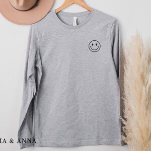 Puede incluir: Una camiseta de manga larga gris jaspeado con un gr&aacute;fico de cara sonriente negra en el pecho izquierdo. La camiseta est&aacute; colgada de una percha de madera. La marca "EMMA & ANNA" est&aacute; impresa en la parte inferior.
