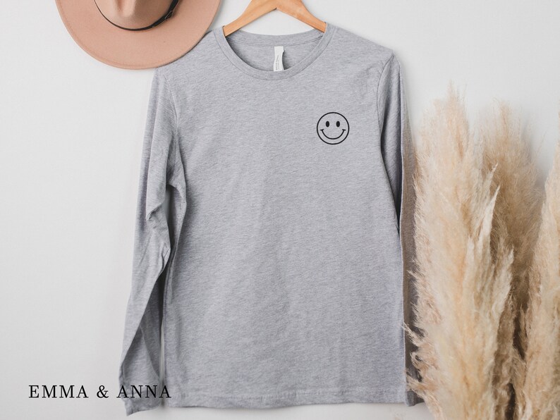 Puede incluir: Una camiseta de manga larga gris jaspeado con un gr&aacute;fico de cara sonriente negra en el pecho izquierdo. La camiseta est&aacute; colgada de una percha de madera. La marca "EMMA & ANNA" est&aacute; impresa en la parte inferior.