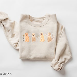 May include: A beige sweatshirt with a graphic of four golden retriever dogs sitting in a row with wildflowers behind them.