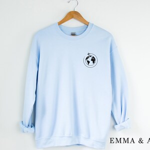 May include: A light blue sweatshirt with a black graphic of a globe with a plane flying around it. The text "EMMA & ANNA" is printed on the bottom right corner of the image.