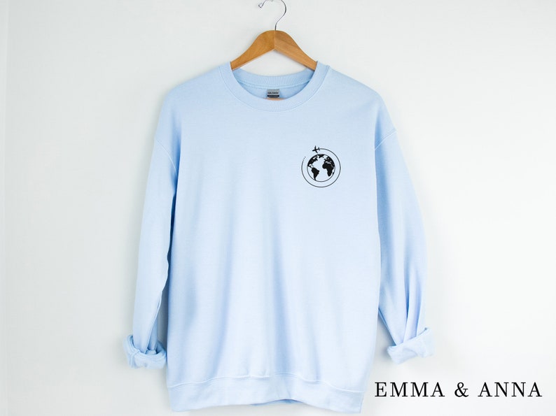 May include: A light blue sweatshirt with a black graphic of a globe with a plane flying around it. The text "EMMA & ANNA" is printed on the bottom right corner of the image.