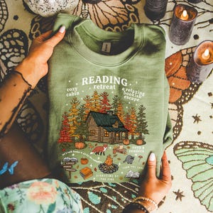 Op de afbeelding: Groen sweatshirt met een afbeelding van een hut in het bos, pompoenen, een vos en de tekst "Reading Retreat". Het ontwerp bevat de zinnen "cozy cabin", "a relaxing bookish escape" en "storytime by the fire".