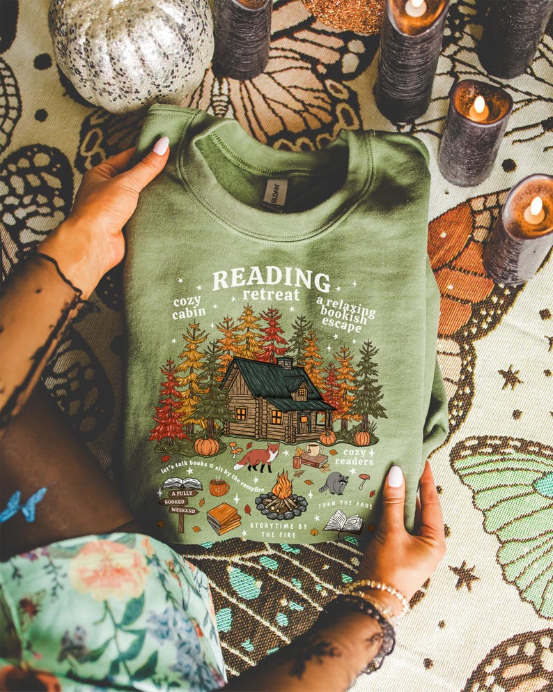 Op de afbeelding: Groen sweatshirt met een afbeelding van een hut in het bos, pompoenen, een vos en de tekst "Reading Retreat". Het ontwerp bevat de zinnen "cozy cabin", "a relaxing bookish escape" en "storytime by the fire".