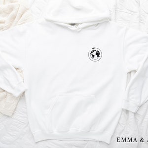 May include: White hoodie with a black graphic of a globe with a plane flying around it. The text "EMMA & ANNA" is printed on the bottom right corner of the image.